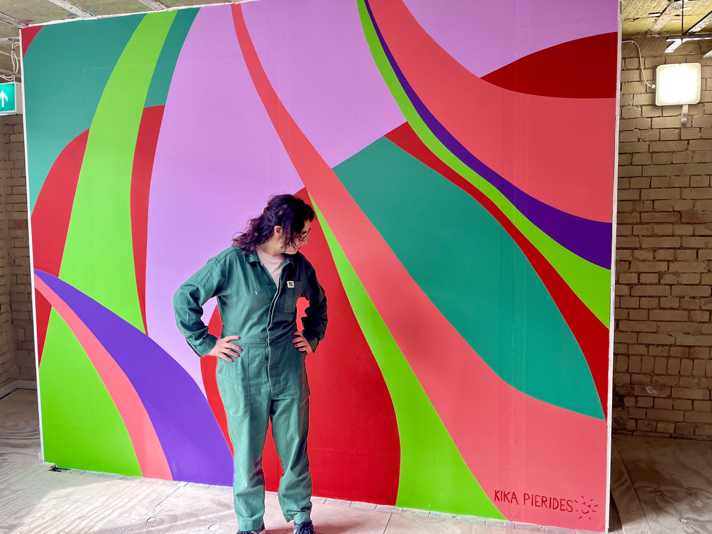 Contemporary patternist Kika Pierides standing in front of her large, vibrant abstract mural with sweeping shapes in pink, green, red and purple