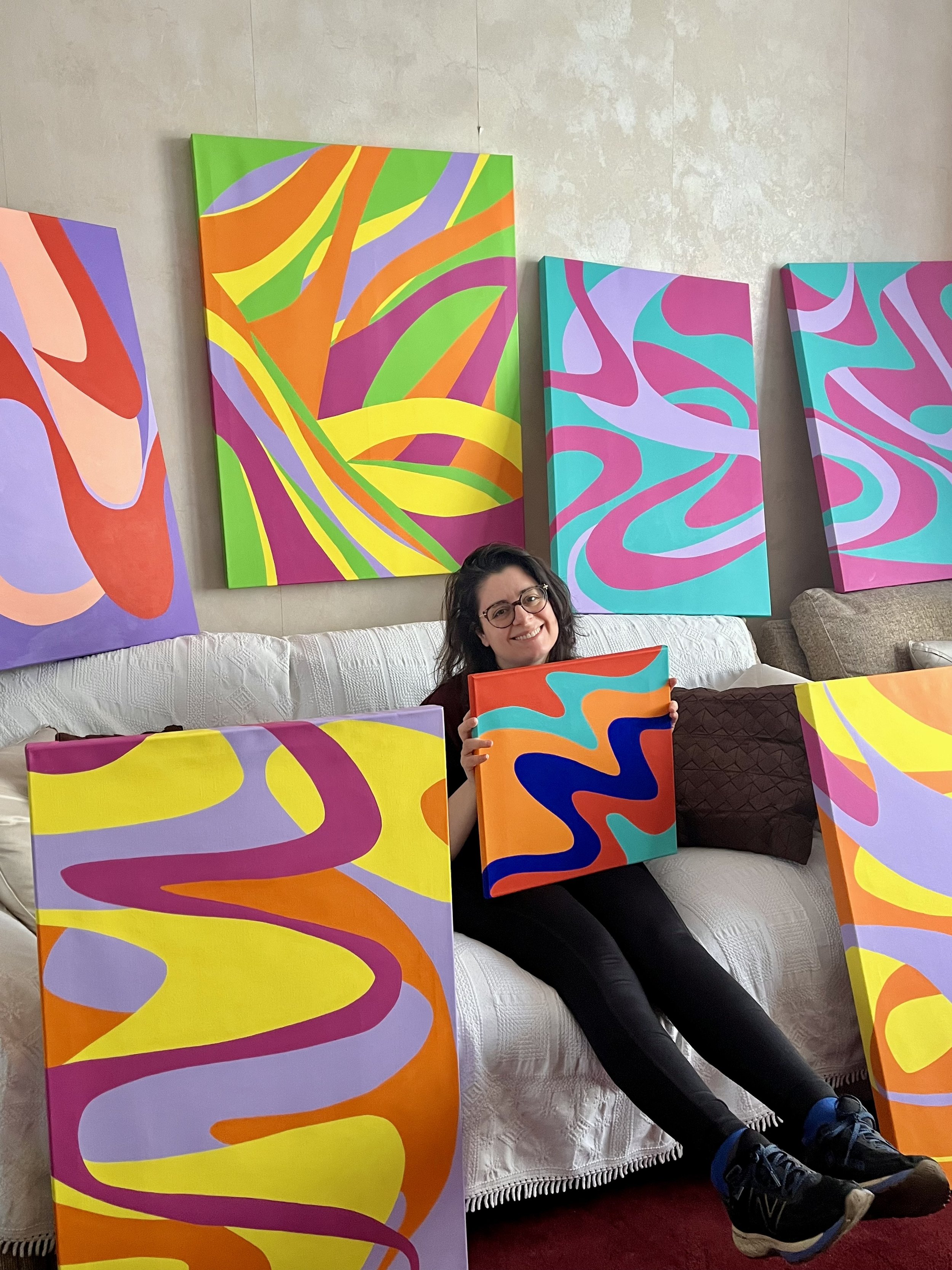 Contemporary patternist and textile artist Kika Pierides sitting on a couch, holding a colourful abstract painting, with more paintings leaning against the couch and hanging on the wall behind her